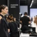 Behind-the-scenes fashion week scene showing a model being prepared backstage beside clothing racks, stylists, and a runway show.